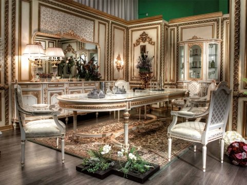 This picture shows a luxurious living room with modern design elements. The walls are painted white, and there is a stylish ivory colored armchair in the center of the room. The floor is a dark-toned parquet, and the fireplace is decorated with marble. The walls are decorated with tall mirrors, and there is a large abstract painting above the sofa. On either side of the armchair, there are small round side tables with lamps on top.