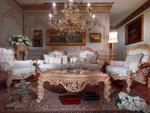 This picture shows a modern living room that features an extravagant crystal chandelier. The furniture is lavish and comfortable, with refined upholstery and a leather armchair. The walls are decorated with a large wall mirror framed in gold and a vibrant abstract wall art. To complete the design, the floor is covered in luxurious, warm-colored carpets.