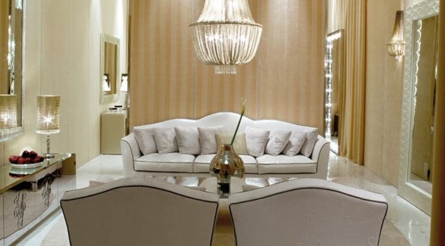 This picture shows a modern, luxurious living room. The walls of the room have been decorated with cream-colored wallpaper and the floor is covered with a white rug, giving the room a soft and airy feeling. There is a large sofa in the center that is upholstered in a light pink fabric with matching cushions, and it is surrounded by several chairs of different colors and styles. There is an elegant coffee table in the middle of the room, and on each side of it are tall standing lamps. The entire room is decorated with art and framed photographs, creating a warm