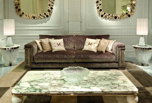 This picture shows an opulent living room in a modern home. The room features a large sectional sofa upholstered in a luxurious gold fabric with coordinating throw pillows. The furniture is set against an elegant marble-tiled wall and is accessorized with a mirrored glass coffee table and two gold side tables with lamps. The ceiling features a crystal chandelier that adds additional sparkle to the space.