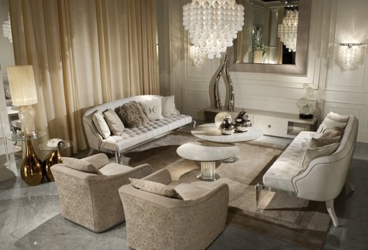 This is a picture of a modern, luxurious living room. It features a white couch with colorful pillows and throws, a glass coffee table, a large abstract painting on the wall, and a white rug. There is a modern chandelier suspended from the ceiling and a unique, curvy side table. The room also includes several large windows that allow natural light to stream in from outside.