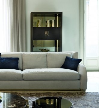 This is a photo of a modern, luxurious living room with an espresso-colored sectional sofa, a marble top coffee table, and a contemporary rug. The walls have an ornate gold trim and sconces casting a warm light on the space. The room also features a dining area with a large glass table and four curved chairs upholstered in luxurious fabric. A wooden console table is also seen in the background, featuring vases and a lamp.