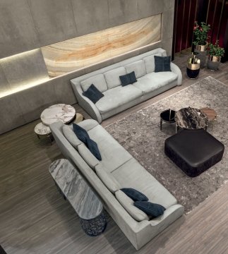 This picture shows a modern master bedroom suite with luxury furniture. The room has a large bed set in front of a floor-to-ceiling window, which looks out onto a balcony with a skyline view. The walls are decorated with textured wallpaper and metallic accents while the floor is covered with a grey rug. The luxurious furniture includes a mirrored dresser, an armchair upholstered with a dark patterned fabric, a wall sconce, and a side table. The room also includes a modern chandelier hanging from the ceiling.