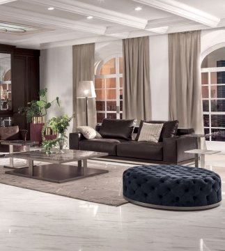 The picture shows a well-decorated, luxurious living room. The room features beige walls, a white ceiling with cove molding, and multiple large windows covered by golden curtains. A modern dark grey sofa is highlighted in the center of the room, flanked by two matching armchairs. A large, round coffee table sits between them, with a gray and golden area rug beneath. The room is completed by a wooden sideboard against the wall, and various pieces of art and light fixtures throughout.