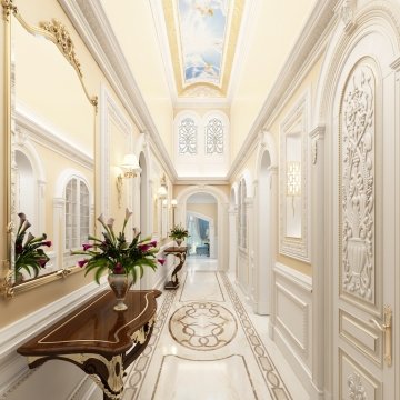 The picture shows a grand entrance hallway that has been designed with luxurious and modern decor. The walls are lined with a marble pattern and there are two large columns on either side of the doorway. There is also a crystal chandelier hanging from the ceiling and several comfortable armchairs set up in the room. On the left side of the entrance there is an elegant mahogany table and two grey armchairs, and a white vase with flowers sitting on top.