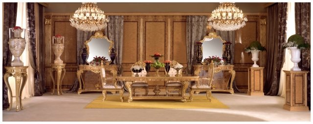 The picture shows a luxurious bedroom, decorated in a classic and elegant style. On the left side is a white bed with an ornate wooden headboard. The bed is dressed with crisp white linens, and there is a large, silky white throw blanket draped over it. The walls are painted a soft beige color, and there is a large mirrored dresser on the right side of the room. Above the dresser is an ornate oil painting, and the floor is covered in a beige rug. A small lounge chair is on the far side of the room, and