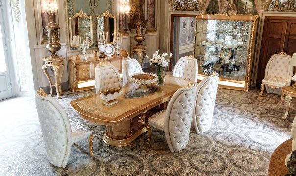 This picture shows a grand and elegant dining room. The room features a long marble dining table with intricate gold accents along the border as well as detailed carved legs. There are eight upholstered chairs around the table, featuring a light pink color with lush velvet fabric. The walls of the room are covered with cream wallpaper with a flower and branch pattern, and a golden chandelier hangs from the ceiling. The room is finished with a Persian rug in the middle of the floor, adding a touch of warmth to the area.