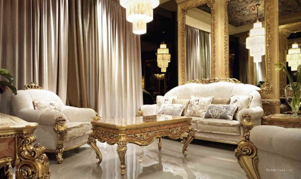 This photograph shows a modern living room with an elegant white and grey color scheme. The walls are adorned with framed art pieces and feature recessed lighting for ambient mood lighting. The room is furnished with a large grey upholstered sofa, two armchairs in the same style, two accent chairs with light-wood frames and a glass coffee table with stylized legs. An eye-catching focal point is the fireplace with a white marble surround and a wall of cabinets for storage above it.