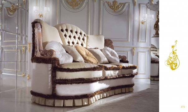 This picture shows a high-end, modern bedroom suite with luxurious furniture and fixtures. The room has a wood-paneled ceiling and walls, with a large window overlooking a beautiful garden. The bed is upholstered in white leather, with a matching armchair and ottoman. A patterned gold and cream area rug covers the floor beneath them. On either side of the bed are two tall mirrored nightstands and lamps. A chandelier hangs from the ceiling, and a small chest of drawers sits against one wall.