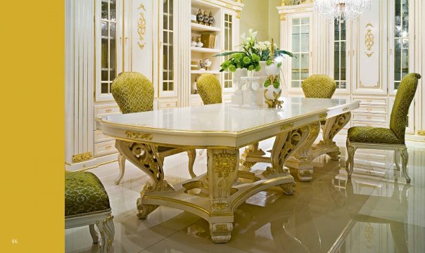 This picture shows a luxurious modern bedroom design. It features a white tufted headboard with a mirrored bedframe, two nightstands on either side, and decorative lamps. The room is decorated with an ornate gold chandelier and wall panels with an intricate pattern. The walls are painted in a soft grey hue, and the floors are covered in plush white carpeting. There is a large window at the foot of the bed to let in natural light, and the space is finished off with a few pieces of modern furniture.