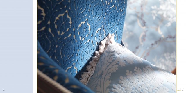 This picture is of a small, white kitten perched on the arm of a gray loveseat. The kitten is curled up with its head resting on its paws, looking off into the distance. Its eyes are closed, suggesting that it may be sleeping and dreaming.