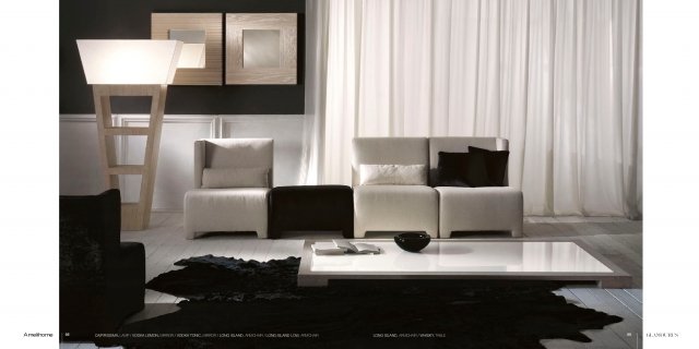 This picture shows a luxurious modern living room with a contemporary design. The room features a white sofa set with a black and gold ottoman in the center, an ivory marble coffee table, and a round golden mirror on the wall. A white chandelier hangs from the ceiling, while two white ceramic floor lamps provide additional lighting. The walls are painted in a light gray hue and decorated with abstract art pieces.