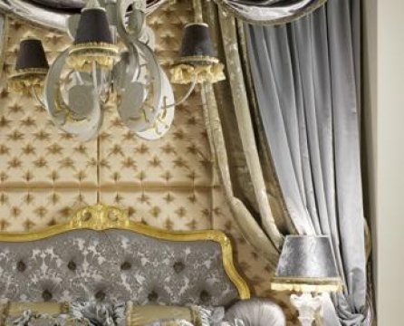 This picture shows a luxurious living room, featuring two red velvet sofas arranged around a marble-topped coffee table. On either side of the sofas are silver-colored armchairs with gold gilded details. There is a large fireplace in the corner with a mirror above it and a set of white shelves displaying various objects. The walls are covered in a beige wallpaper with intricate patterns and the floor is covered in a dark brown wooden parquet. Various lamps, including a large crystal chandelier, are suspended from the ceiling, lighting up the room.