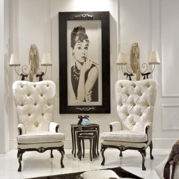 The picture shows a living room with a contemporary Art-Deco style. The walls are painted in a deep blue color and the furniture is upholstered in a light grey velvet fabric. There is a large, modern chandelier hanging from the ceiling that gives the room an elegant and luxurious feel. The floor is covered in a beige carpet and there is a large modern couch in the center of the room.