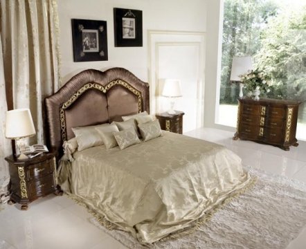 This picture shows a luxurious living space decorated with a contemporary style. The room features a large, off-white sofa paired with a plush, white armchair and two black accent chairs. There is a large, wooden coffee table in the center of the room and an area rug with a geometric pattern underneath. The walls are painted light gray and are adorned with artwork and silver wall fixtures. Other details include a stone fireplace, dark wood sideboard, and glass lamps.