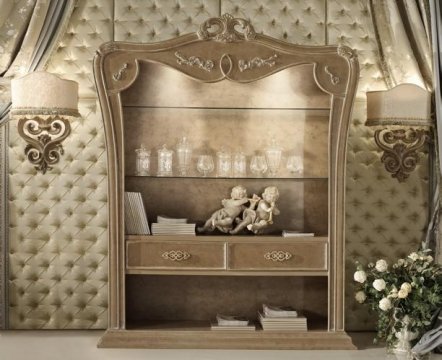 This picture shows a luxurious bedroom in an elegant style. The room is decorated with ornate, gold furniture and accents, including the large headboard on the canopy bed, which is draped in deep red fabric. The walls are covered in a light, beige wallpaper with a subtle, dark blue pattern. On the wall to the right is a large window, framed by heavy, velvet curtains. The floor is covered in a thick, off-white carpet, and there is a pair of comfortable armchairs in the corner.