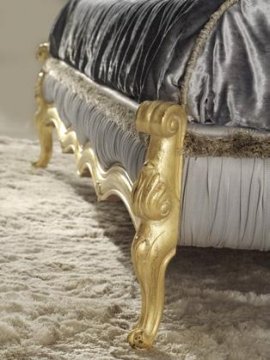 This is a picture of a luxurious bedroom with white and gold accents. It has a large, cushioned headboard with intricate gold details and four tall gold-painted wooden posts. The walls are in a light gray shade with a shiny gold pattern resembling a sunburst. The bed is covered with a duvet in a pale peach hue, and the bedspread is in a white hue with a ruffled texture. The room features several pieces of modern furniture in shades of white, such as a tufted armchair, a small round table, and an accent table. Several white