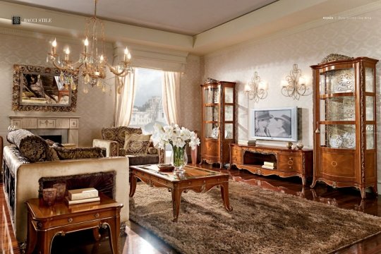 This picture shows a luxurious living room with a beautiful white and gold theme. The room features a large, comfortable ivory sofa and two matching armchairs upholstered in white and gold fabric. A light wood coffee table is situated in front of the sofa. The walls are painted a light shade of gray and decorated with ornate mirrors and a large gold-framed portrait. A large grey rug covers most of the floor, providing an elegant backdrop for the furniture in the room.