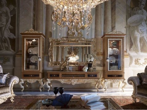 This picture shows an opulent, modern bathroom. It features a black marble floor and walls that are lined with a gold-toned mosaic tile. The room is brightly lit by the white tub in the center of the room, which is equipped with a gold-plated faucet. In addition, there is a vanity with a gold-plated sink, two gilded light fixtures, a large framed mirror, and several vases.