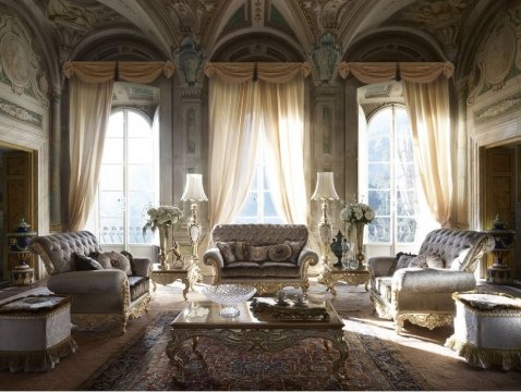 This picture shows a luxurious living room with modern design elements. The walls are painted in a grey-blue tone, creating a calming atmosphere. The walls are adorned with wall art, adding to the modern feel. The large cream couch provides ample seating for guests, and the decorative pillows add a touch of color and style. A glass coffee table is placed in the center of the room, surrounded by cushioned armchairs. The space is illuminated by several hanging light fixtures, as well as floor lamps.