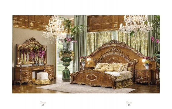 This picture shows a modern, luxurious bedroom with a balcony. The room appears to be well lit, with warm colors and gold accents. The bedroom features a round velvet headboard, two velvet armchairs, and a large floor-to-ceiling window overlooking a balcony. A chandelier hangs overhead, adding an elegant touch to the room. The bed is covered in white and light gray linens, and there is an upholstered footstool next to the armchair.