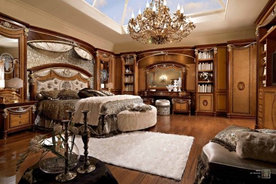 This picture appears to show a luxurious master bedroom suite with a rounded bed frame, an oval-shaped headboard upholstered with a rich, textured fabric, and two nightstands with glass-top surfaces. Adorning the walls is a modern chandelier, a large mirror, and a framed art piece. The floor is covered in a soft, neutral-colored rug and the room is decorated with several ornate throw pillows.