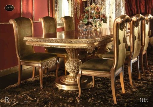 This image shows a large marble-tiled bathroom. The walls and floor are white marble and the ceiling has a decorative patterned panel of gold circles. The bathroom has two ornate, wooden vanities with white countertops and matching gold wall sconces. At the far end of the room there is a large walk-in shower with glass doors and framed by white marble tiles. There is also a large soaking tub in the center of the room.