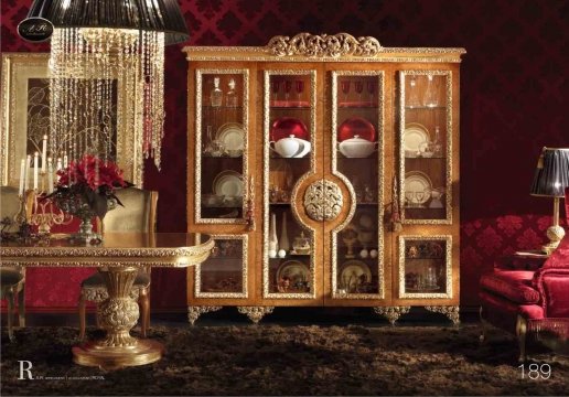 The picture shows an elegant entrance lobby area with a rich, marble floor, and beige walls. There is a wooden half wall with a gold-plated frame that serves as a partition. A gold chandelier hangs from the ceiling, adding a luxurious touch to the room. Additionally, there are two chairs and two tables with a vase of flowers sitting atop each one.