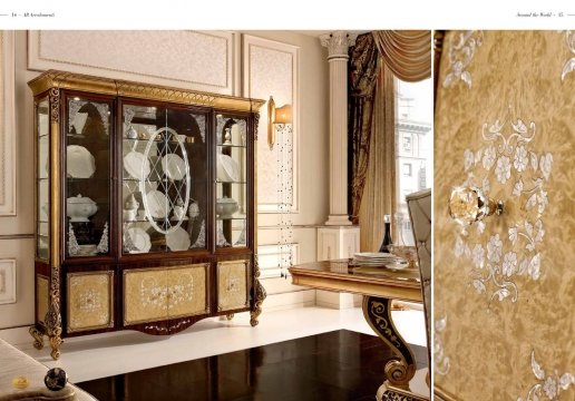 This picture shows a luxurious, modern living room space. The room has a beige and white color scheme with both wallpaper and textured wall panels. A stunning crystal chandelier hangs from the ceiling, framed by a lighted archway above it. The furniture is upholstered in white textiles and features golden accents. In the center of the room, a white coffee table is set upon a grey and brown rug, with a white and gold decorative vase and an ornate framed mirror on top.