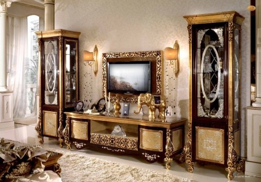 This picture shows a modern living room with a tan couch, an orange and navy blue armchair, and a gray rug. There is a gray coffee table with a vase of flowers in the middle, two white end tables with lamps on either side, and a painting above the couch.