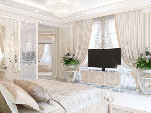 The picture shows a luxurious bedroom with elegant gold accents and decor. The walls are a light beige color, and the furniture is upholstered in cream and gold fabric. There is a low, ornate bed frame covered in a white duvet and pillows, and a tufted armchair and ottoman nearby. On one side of the room is a large window with floor-to-ceiling curtains, and on the other side is a built-in bookcase full of books and other decorative items. The room also features a white chandelier, mirrored