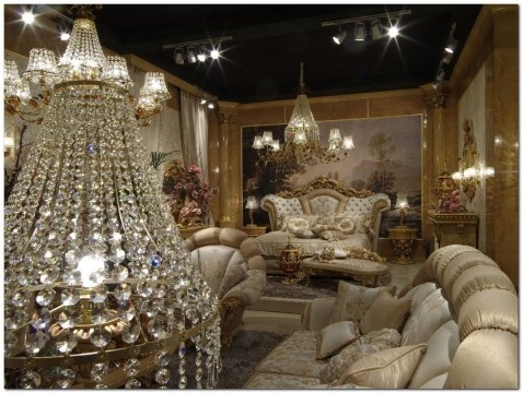 This picture shows an interior design featuring a large, white sofa, two armchairs and a coffee table with a decorative bowl of flowers in the center. The walls are painted a light grey, and the room is lit by a bronze chandelier. A black-and-white rug is placed on the floor, and several pieces of abstract artwork hang on the walls, adding a touch of color.