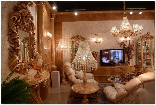 This is a picture of a luxurious living area. It features an ornately decorated white and gold sofa, two armchairs in a similar color scheme, and a tufted rectangular ottoman. The walls are painted a pale blue and the room is accented with detailed crown moulding and white and gold curtains. The two tables to the sides of the seating area each have vases with a bouquet of white roses and various sculptures. The coffee table between them has a large glass top and is surrounded by white and gold books, a lamp, and other decorative accents.