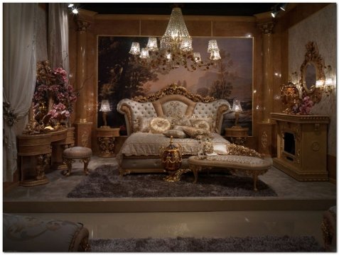 This picture shows a modern living room design featuring luxurious gray and white furniture accents. The walls are painted a light gray and adorned with wall art and abstract sculptures that are either black or dark gray in color. The floor is covered in a plush gray carpet, and the large window provides plenty of natural light for the space. In the center of the room is a round glass table with two comfortable armchairs. On the wall to the left is an entertainment center with a large flat-screen television.