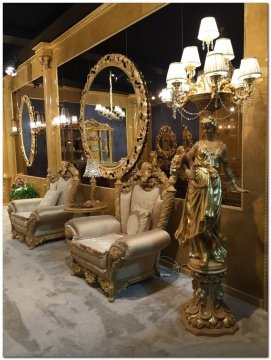 This picture shows a luxurious bedroom interior design featuring a four-poster bed with an ornate golden canopy and matching drapery. The walls are painted in a creamy off-white color and a plush navy blue rug is spread across the floor. Two lounge chairs with white upholstery provide additional seating and a large side table stands in between them with a vase of fresh flowers on top. Other decorative elements such as beautiful mirrors and paintings add to the luxurious atmosphere of the bedroom.