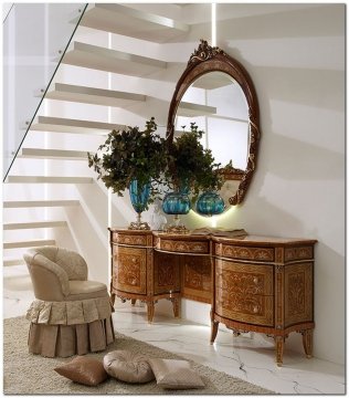 This picture shows a luxurious sitting room with light blue walls, a large stone fireplace, and two beautiful white sofas. The sofas are adorned with golden detail and arranged in an "L" shape around a large glass coffee table. The room is illuminated by several tasteful chandeliers and light fixtures, adding to the regal atmosphere of the space. A large oval mirror is mounted on the wall above the fireplace, reflecting the bright colors of the room and its plush furnishings.