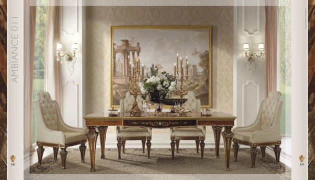 This picture shows an opulent, luxurious hotel suite. It features a large, comfortable seating area with two plush, white sofas, a glass coffee table, and various decor items, such as a floor lamp and a small potted plant. Behind the sofa is a backdrop of large windows, with sheer drapes and a patterned wallpaper. On the right is a paneled door, likely leading to the bedroom. The whole room is decorated in a modern and elegant style, with a creamy color scheme and gold accents.