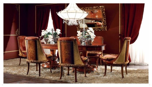 This picture shows a luxurious living room with a large white leather sofa and matching armchairs. The walls and ceiling are painted golden, and there is a large gold-framed mirror on the wall. There is an intricate crystal chandelier above the seating area, and in the corner a classical column is decorated with a luxurious tapestry. A small white table sits between the sofa and armchairs and there is a glass-top coffee table in the center of the room.