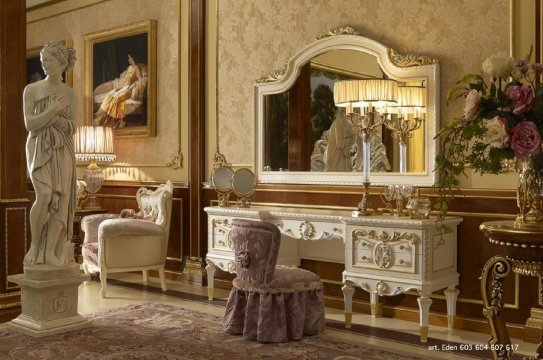 This picture shows a luxurious bedroom with an art deco-style interior design. The walls are light yellow and feature a stucco pattern. The ceiling is ornamented with a beautiful chandelier and light gold accents. The flooring is dark wood with several carpets. The bed has a light blue headboard and footboard, with intricate gold detailing around the edges. There are large curtains framing the windows and a white armchair near the bed.