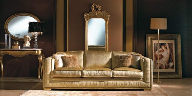 This picture shows a luxurious gold-trimmed living room. It features an ornately-styled fireplace, a white marble floor, and a beige sofa and chairs. A glass and gold coffee table, along with several decorative pillows, provide a modern touch. The curtains on the windows are made of a rich cream fabric, while the decorative vases and wall art are in shades of golden yellow. The chandelier and sconces add an extra sparkle to this elegant living room.