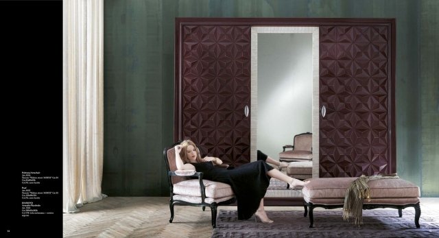 This picture shows a luxurious living room with golden accents. The room is decorated with furniture including an armchair, a sofa, and a glass coffee table. The walls are covered in a muted grey wallpaper with gold designs, and the floor features a white geometric rug. There is a ceiling-mounted chandelier as well as wall sconces around the room. On the walls are various art pieces, including a large painting of a vase against a yellow background.