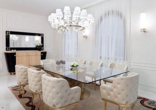 This picture shows a modern living room with a large sectional sofa in a light gray color. There is a round glass-top coffee table in the middle, and two high back chairs in a matching color. The walls are decorated with abstract canvas art pieces, and a large mirror is hung above the sofa. The room has hardwood floors and a large, crystal chandelier hanging from the ceiling.