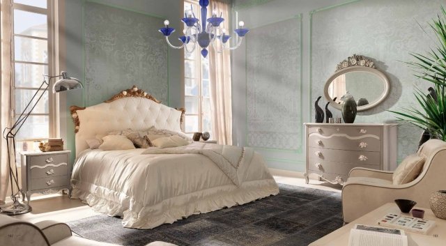 This picture shows a modern, luxurious bedroom. It has a grand, traditional-style bed with a polished gray headboard and white bedding. On the left side of the bed is a beautiful light blue accent chair with white trim and intricate detailing. On the right side is a large nightstand with drawers, and a white modern lamp. The dark gray wall behind it offers a contrast to the other lighter colors in the room, and the light hardwood floors complete the look.