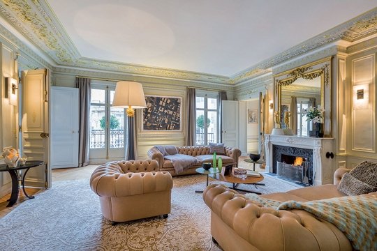 This picture shows a luxurious, modern living room. It features a navy blue velvet sofa with leather trim, two matching ottomans, and two white armchairs. The walls are painted an off-white color, and a beige-colored area rug covers the hardwood floor. A contemporary chandelier hangs from the ceiling, and several framed art pieces are displayed on the walls.