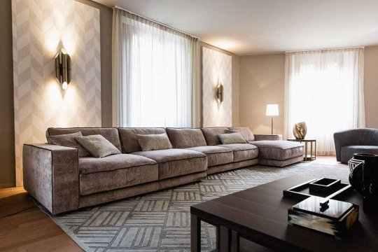 This photo shows a luxurious and modern living room with a white leather sectional sofa and a matching ottoman. There is a beige-colored accent wall with a decorative mirror and two matching bronze sconces on either side. The adjacent wall has a large built-in television with more shelves for storage. Additionally, there is an intricately-patterned area rug in the middle of the room that ties the look together.