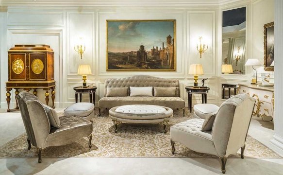 This picture shows a luxurious living room with an ornate black and gold framed mirror hung above the fireplace. The room has a plush white sofa and two chairs facing towards the fireplace, and a large wine table off to the side. The walls are decorated in a deep red wallpaper with gold accents, and two crystal chandeliers hang from the ceiling.