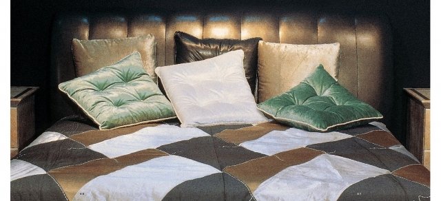 This picture shows a luxurious master bedroom featuring an ornately carved white wood bed frame, with a large tufted headboard and detailed velvet upholstery. On either side of the bed is a pair of glass-shaded lamps and two intricately carved wooden nightstands. The room also features a white silk canopy draped over the bed and a red and gold Persian rug on the floor. High ceilings and a large window provide plenty of natural light, and a marble fireplace with a large mirror hangs on one wall.